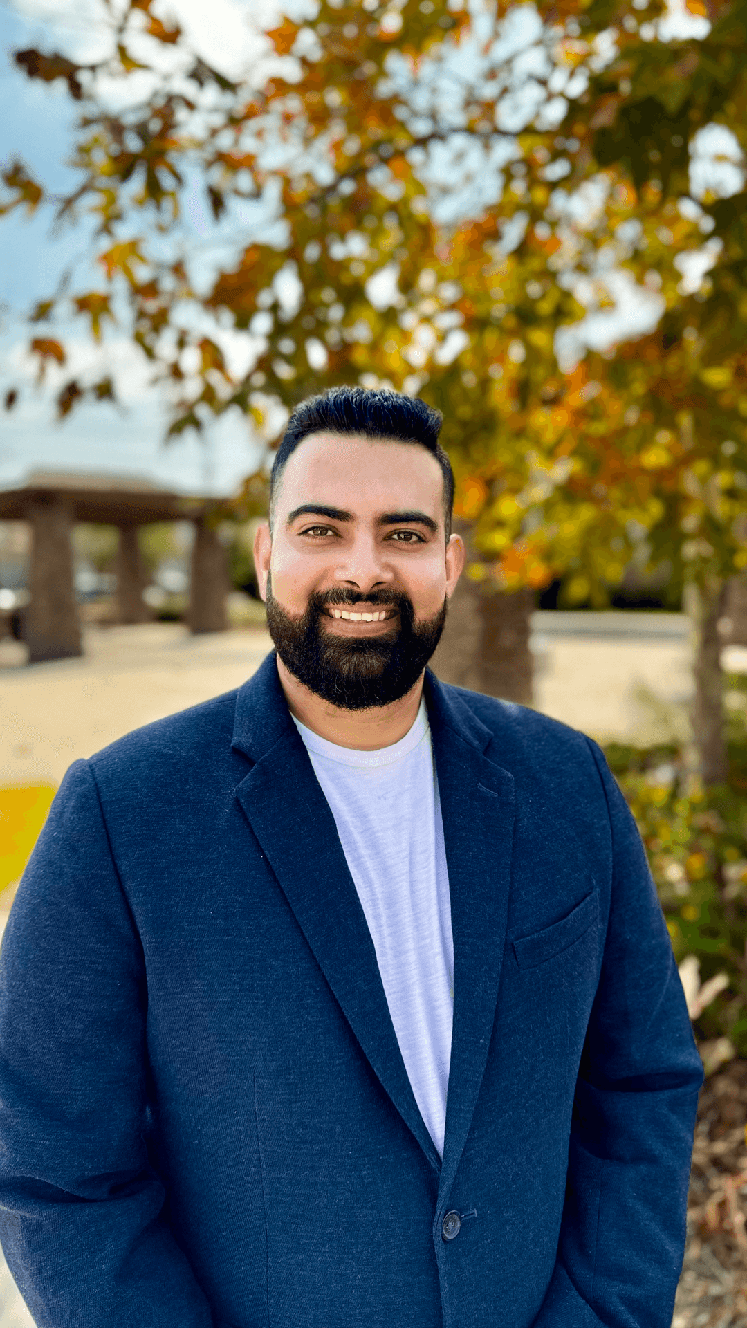 Person in a blue blazer and white shirt standing outdoors with autumn leaves and trees in the background.